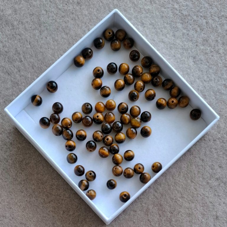 Tigerauge Perlen Braune und schwarze Perlen in einer weißen Schachtel, verstreut auf einem hellen Untergrund.