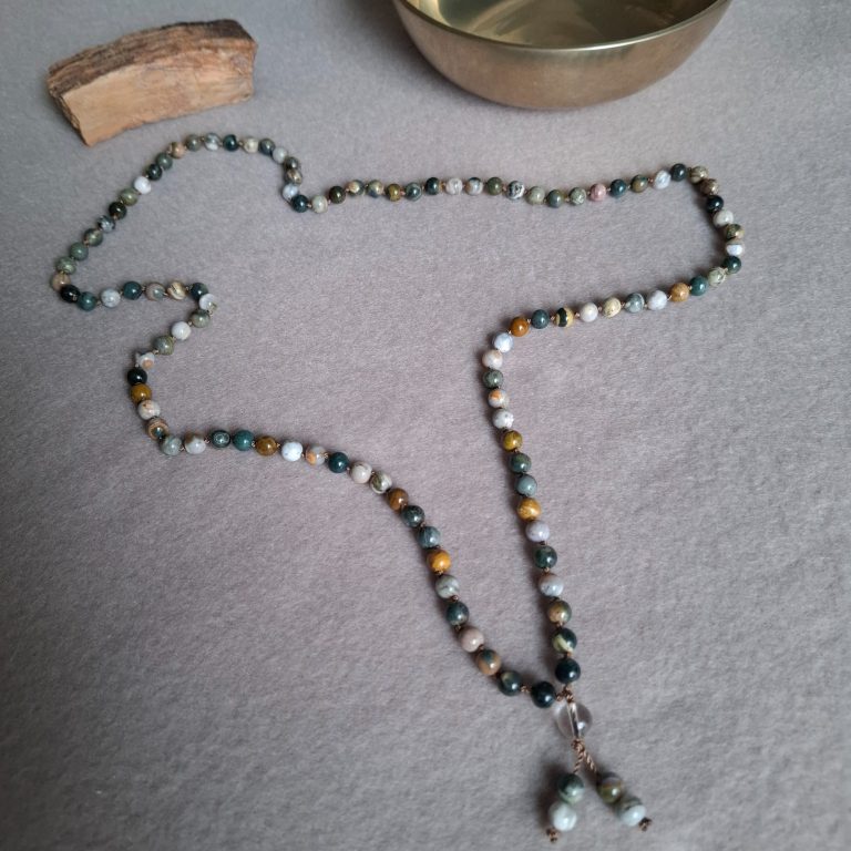 Kette aus bunten Perlen mit einem Anhänger und einer Klangschale und einem Holzstück im Hintergrund.